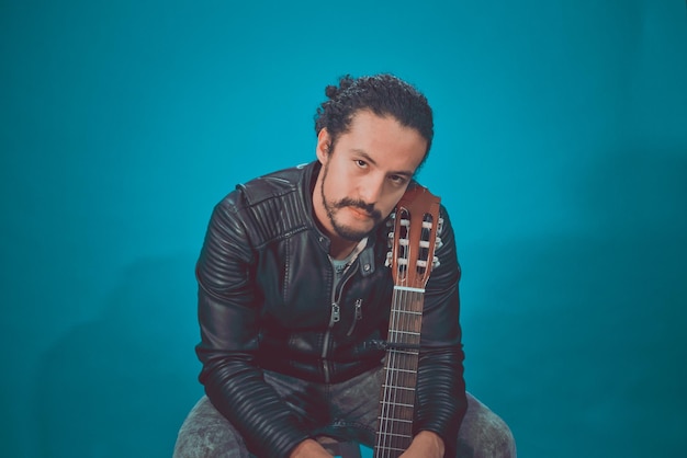 Portrait von attraktiven lateinamerikanischen Jugendlichen in Rockerjacke, auf blauem Hintergrund, mit Akustikgitarre