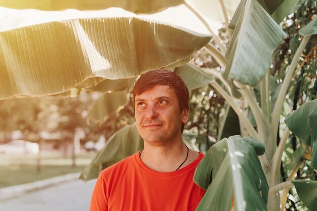 Portrait von Gesicht glücklich lächelnd unrasiert 40 Jahre Mann Alleinreisender mit Stoppeln auf dem Hintergrund von grünen Pflanzen Palmenblättern in der Natur an sonnigen Sommertagen auf Urlaubsreisen