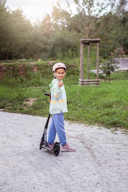 Portrait von niedlichen kleinen kaukasischen Schulmädchen mit Helm genießen Spaß beim Rollerfahren auf asphaltiertem TrackxAin Street Park im Freien an einem sonnigen Tag Gesunde Sportaktivitäten für Kinder draußen