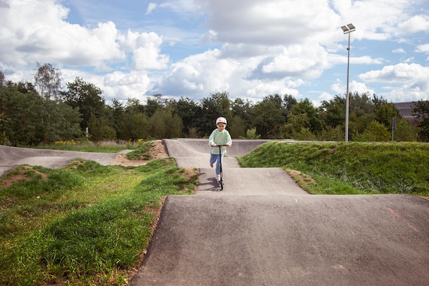 Portrait von niedlichen kleinen kaukasischen Schulmädchen mit Helm genießen Spaß beim Rollerfahren auf asphaltiertem TrackxAin Street Park im Freien an einem sonnigen Tag Gesunde Sportaktivitäten für Kinder draußen