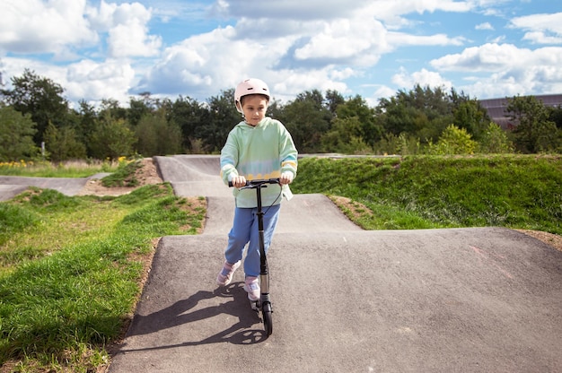 Foto portrait von niedlichen kleinen kaukasischen schulmädchen mit helm genießen spaß beim rollerfahren auf asphaltiertem trackxain street park im freien an einem sonnigen tag gesunde sportaktivitäten für kinder draußen