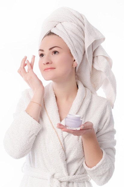 Portret de uma jovem linda em um roupão branco e com uma toalha branca na cabeça aplica creme no rosto com o dedo. Isolado