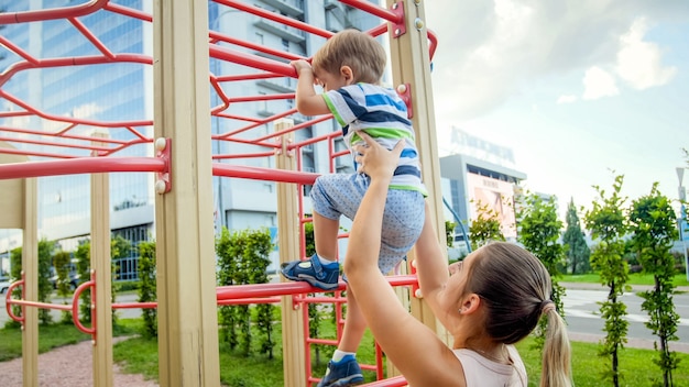 Portriat de uma jovem mãe apoiando e segurando seu filho de 3 anos na escada de metal no parquinho infantil no parque