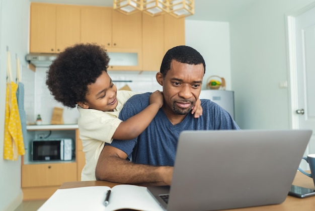 Portriat de un padre afroamericano ocupado que trabaja desde la oficina en casa usando una computadora portátil sentado en la mesa de la cocina con un lindo hijo pequeño jugando casi para molestar a un padre agotado con un niño hiperactivo