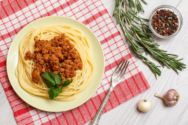 Porzellanteller mit Spaghetti und Bolognese-Sauce, Gabel, Rosmarin und Glasschüssel mit Pimentpaprika auf einer Küchenserviette. Ansicht von oben.