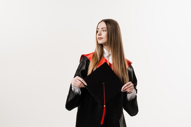 Pós-graduação em vestido de formatura preto está segurando boné nas mãos sobre fundo branco Jovem atraente se formou na faculdade