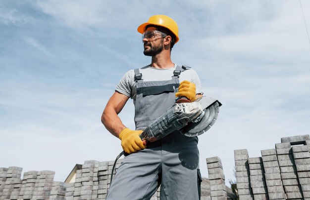 Posando para una cámara con sierra circular Trabajador masculino en uniforme de color amarillo tiene trabajo con pavimento