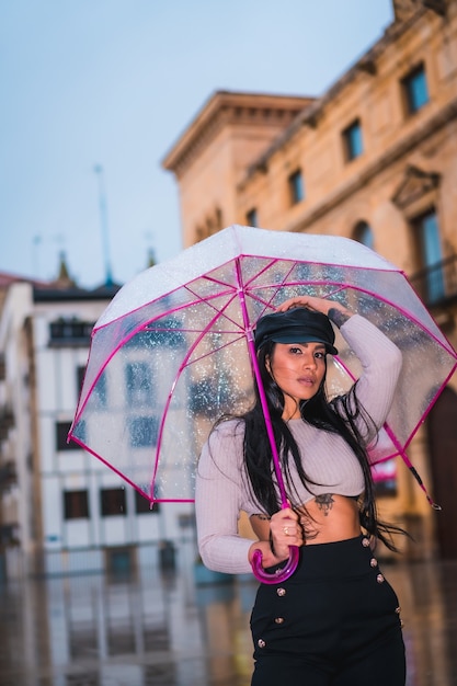 Posando de uma jovem morena latina com um boné de couro na chuva de outono com um guarda-chuva transparente