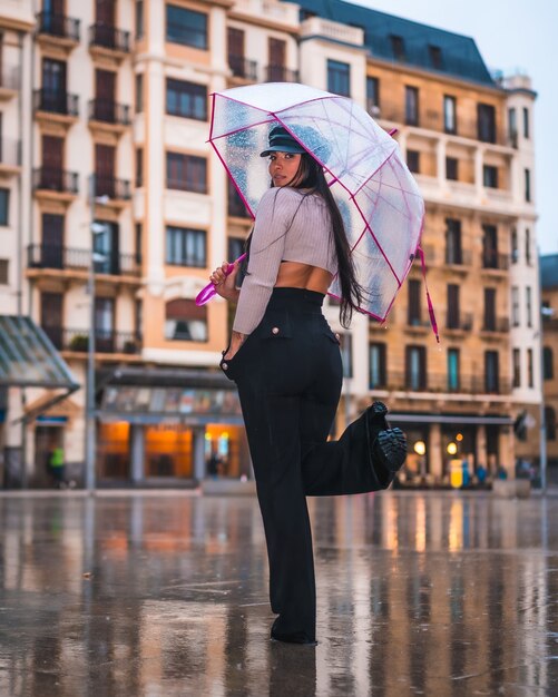 Posando de uma jovem morena latina com um chapéu de couro e calça preta na chuva de outono com um guarda-chuva transparente na cidade