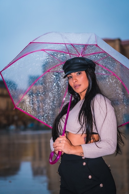 Posando de uma jovem morena latina com um chapéu de couro na chuva com um guarda-chuva transparente