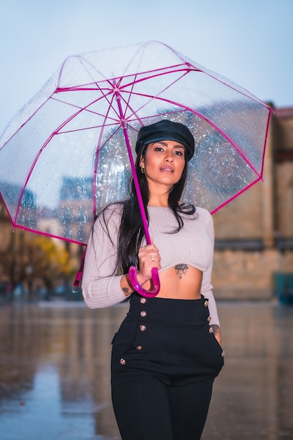 Posando de una joven latina morena con gorra de cuero bajo la lluvia de la ciudad con un paraguas transparente