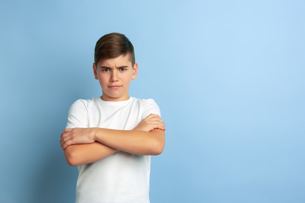 Posando con las manos cruzadas. Retrato de niño caucásico aislado sobre fondo azul de estudio. Modelo masculino adolescente hermoso en la presentación de la camisa blanca. Concepto de emociones humanas, expresión facial, ventas, publicidad.