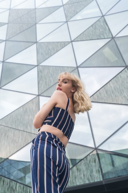 Posando na moda de uma jovem loira em um prédio de vidro, loira em um terno listrado azul, braços para cima, foto vertical