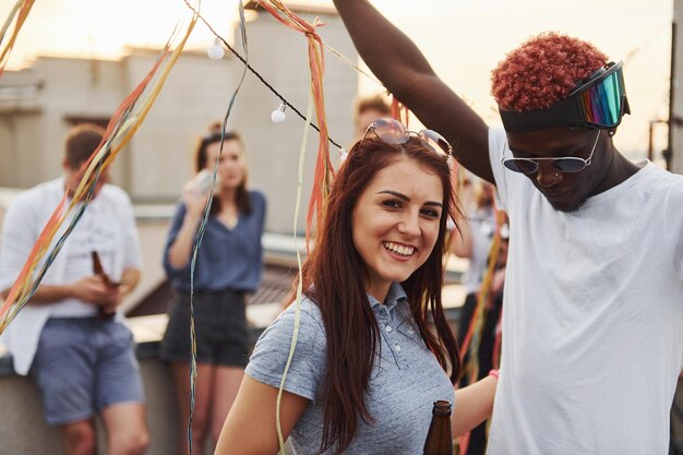 Posando para uma câmera Grupo de jovens em roupas casuais fazem uma festa no telhado juntos durante o dia