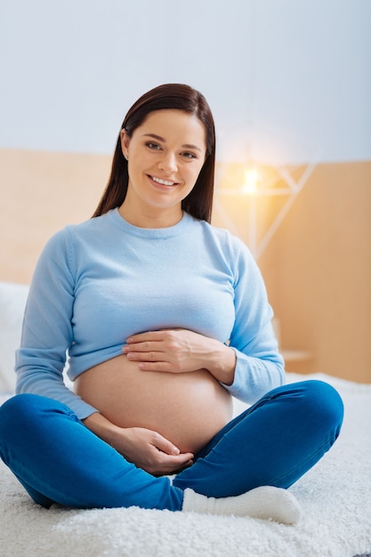 Pose confortável. A futura mamãe encantadora com um sorriso no rosto e as pernas cruzadas