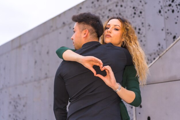 Pose elegante de um casal caucasiano de terno azul e vestido verde, abraçando e fazendo um coração com as mãos