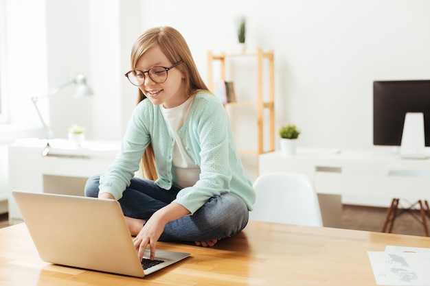 Pose inesperada. Chica inteligente responsable dedicada sentada en la mesa y usando su computadora portátil