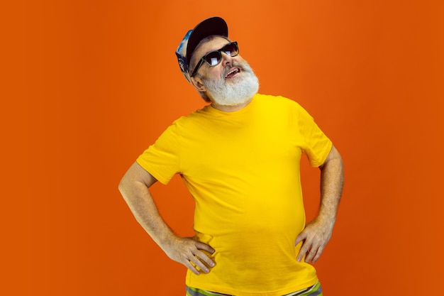 Poses deslumbrantes. Retrato de homem hippie sênior em óculos isolado em fundo laranja do estúdio. Tecnologia e conceito de estilo de vida idoso alegre. Cores da moda, juventude para sempre. Copyspace para seu anúncio.