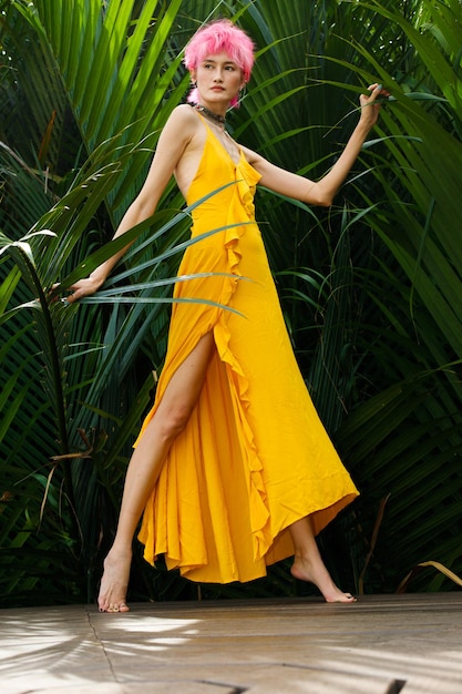 Las poses de moda de la mujer de pelo rosa que muere usan un vestido amarillo y se ven fuertes sobre las hojas verdes tropicales. Asian Slim Female stand elegancia como tiro de moda en concepto Summer tropical zone country