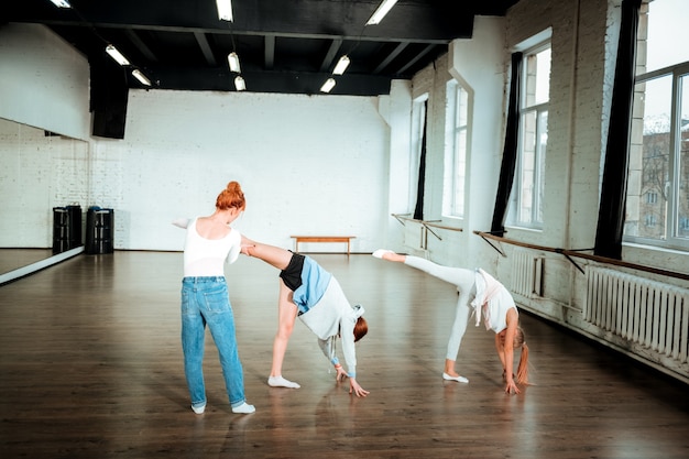 Posicion correcta. Profesora de baile pelirroja vistiendo blue jeans corrigiendo la posición de las piernas de su alumno