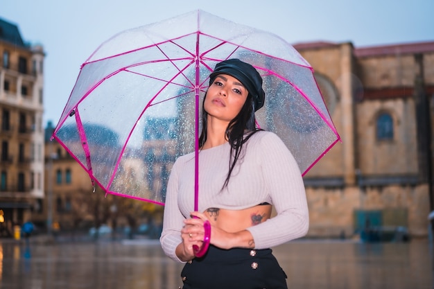 Posieren einer jungen brünetten Latina im Regen der Stadt mit einem durchsichtigen Regenschirm