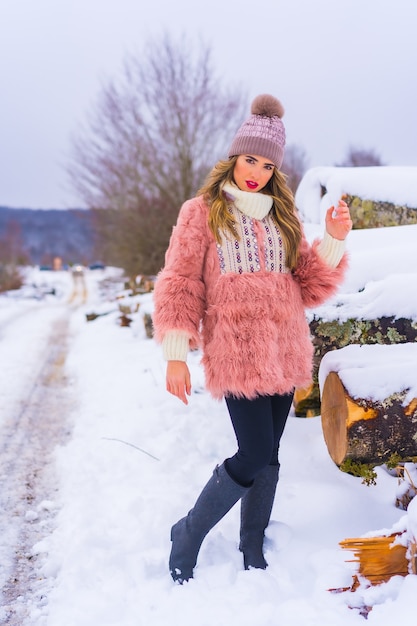 Posing blondes Modell mit rosa Pelzjacke und lila Hut im Schnee. Neben einigen eisgeschnittenen Bäumen auf einem Fußweg winterlicher Lebensstil