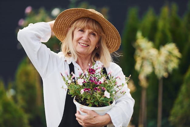 Posing with pot of flowers in hands Senior Woman ist tagsüber im Garten Konzeption von Pflanzen und Jahreszeiten