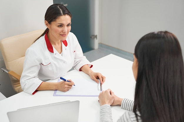 Positiv begeisterte junge Frau, die während der Arbeit in der Klinik Uniform trägt und ihren Patienten während des Termins berät