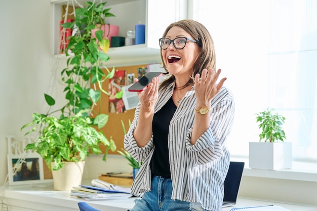 Positiv lächelnde reife Frau, die zu Hause eine Sprachnachricht am Telefon aufzeichnet