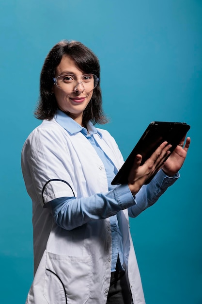 Positiv lächelnder Chemieexperte mit modernem Tablet auf blauem Hintergrund, während er eine Schutzbrille trägt. Fröhlicher Biochemiker mit elektronischem Touchscreen-Gerät.