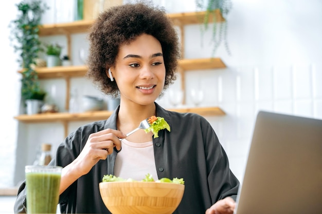 Foto positiv lächelndes afroamerikanisches mädchen in stilvollen kleidern, freelancer, das aus der ferne in einem laptop arbeitet, am tisch sitzt, frisches gemüsesalat isst, während des workflows auf den laptop-bildschirm schaut