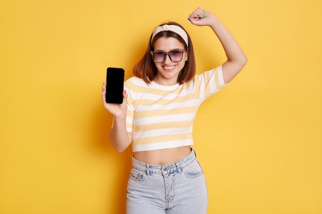 Positiv zufriedene Frau mit Sonnenbrille, gestreiftem T-Shirt und Haarband, geballte Faust, feiert Siegeswetten und gewinnt, zeigt Telefon mit Display, das isoliert über gelbem Hintergrund posiert