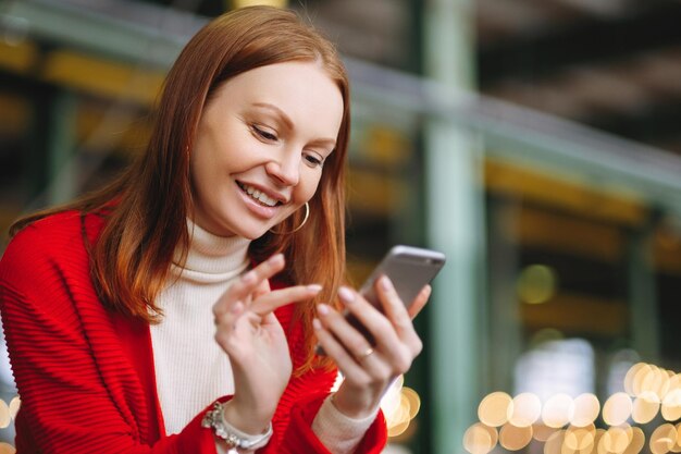 Positiv zufriedenes weibliches Modell verwendet Handydaten verwendet Chat-Anwendung hält modernes Handylächeln positiv in roten Mantel gekleidet wartet auf Antwort oder Anruf von Freund hat braune Haare