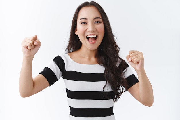 Positiva e entusiástica menina asiática torcendo pelo time, apertando os punhos e sorrindo, encoraja alguém a fazer o seu melhor, sorrindo e apoiando em pé alegre, triunfando sobre o fundo branco