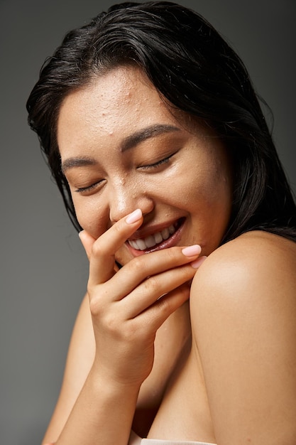 positiva e jovem mulher asiática com cabelo castanho e pele propensa a acne rindo em fundo cinza