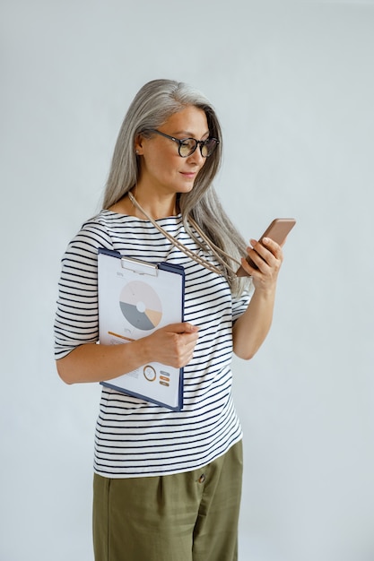 Positiva empresaria asiática madura en ropa casual tiene diagramas y teléfono móvil de pie sobre fondo gris claro en estudio