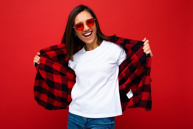 Positiva feliz sonriente joven morena en camiseta blanca casual para maqueta, elegante camisa a cuadros rojos y gafas de sol rojas de moda aisladas sobre fondo rojo con espacio de copia.