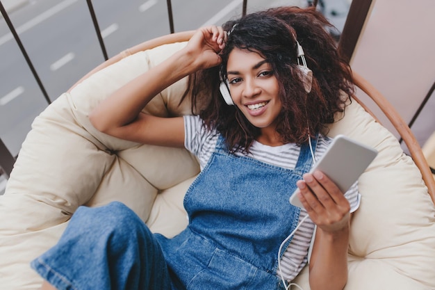 Positiva garota negra com grandes olhos lindos relaxando no terraço e ouvindo música favorita na manhã de verão. Retrato de uma jovem satisfeita com cabelo encaracolado escuro, segurando o smartphone e sorrindo.