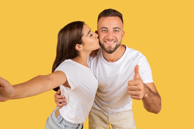 Positiva joven dama caucásica en camiseta blanca beso hombre en cheque divertirse disfrutar de la fecha mostrar pulgar hacia arriba