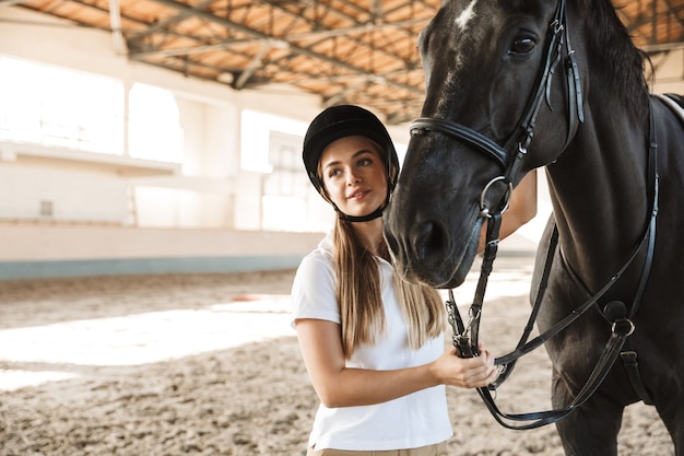 positiva otimista jovem loira linda mulher usando chapéu com cavalo na zona rural em curral na área de treinamento.