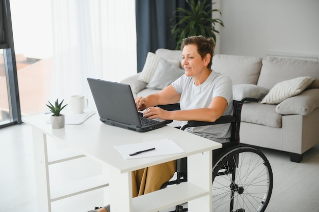 Positive ältere Frau im Rollstuhl, die online an Laptop-IT-Freiberuflern arbeitet