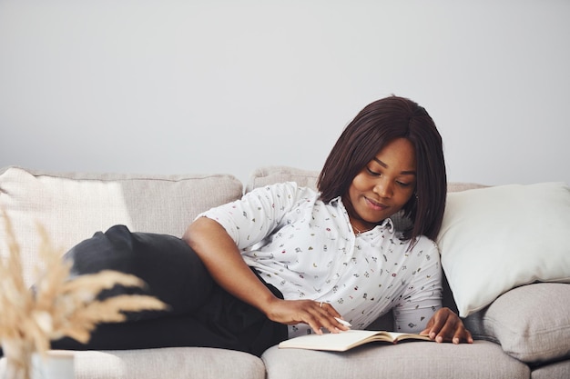 Positive afrikanisch-amerikanische Frau in weißem Hemd, die sich mit Notizblock in den Händen auf das Sofa legt