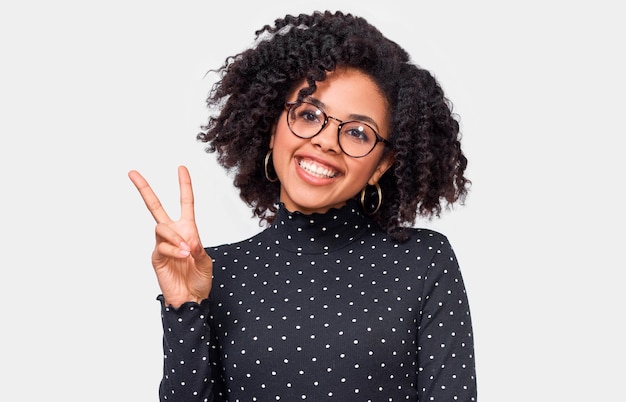 Positive afroamerikanische junge Frau, die breit lächelt und Friedensgeste zeigt, während sie auf die Kamera blickt, die vor weißem Studiohintergrund steht