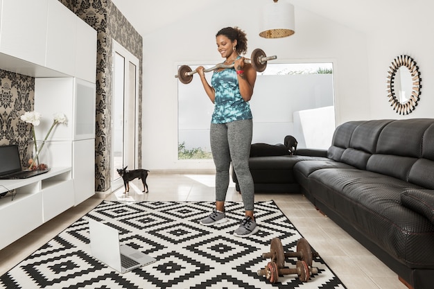 Positive afroamerikanische Sportlerin in Activewear, die Langhantel-Curl-Übungen macht und während des Trainings im Wohnzimmer mit Hunden Online-Videos auf dem Laptop ansieht