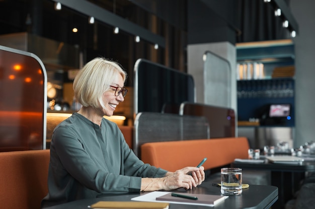 Positive attraktive alte Frau mit Brille, die in einem modischen Café am Tisch sitzt und das Smartphone benutzt, während sie Artikel im mobilen Internet liest