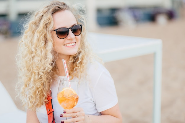 Positive blonde Frau mit dem gelockten Haar, trägt Sonnenbrille, schaut mit glücklichem Ausdruck.