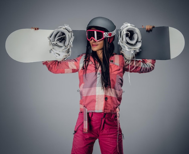 Positive brünette Frau, die ein Snowboard auf ihrer Schulter in einem Studio auf grauem Hintergrund hält