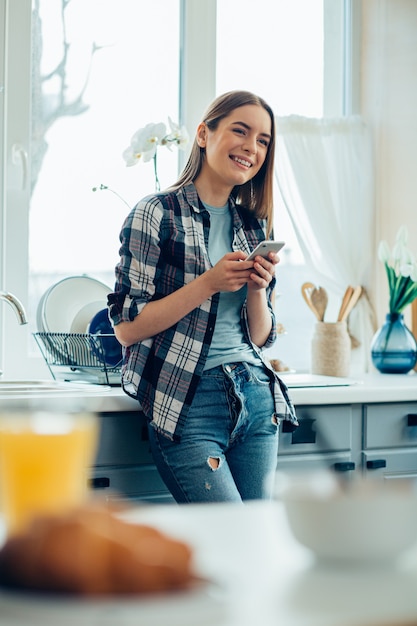Positive Dame in Freizeitkleidung, die ein Smartphone hält, während sie in der Küche steht