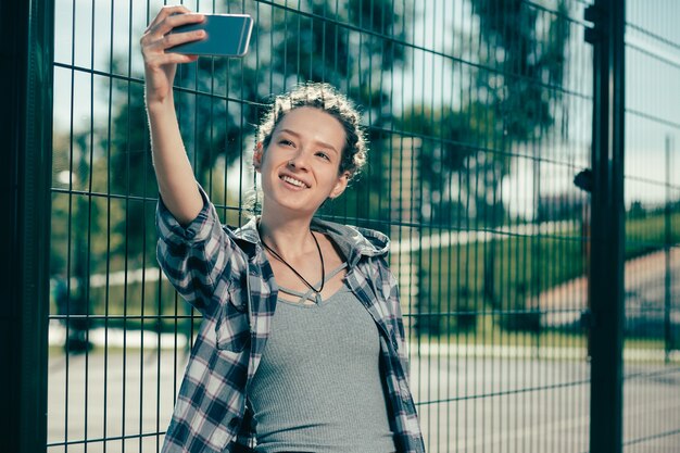 Positive Dame mit Dreadlocks, die lächeln und in der Nähe des Maschendrahtzauns stehen, während sie Selfies machen