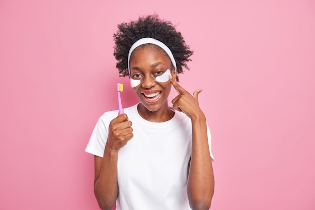 Positive dunkelhäutige Frau mit natürlichem lockigem Haar trägt Schönheitspads unter den Augen auf, hält Zahnbürste durchläuft tägliche hygienische Routine